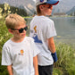 Two boys wearing white graphic t-shirts stand outdoors by the Arosa Obersee lake with mountains in the background. The boy on the right wears a blue and white cap and sunglasses, while the boy on the left wears black sunglasses.