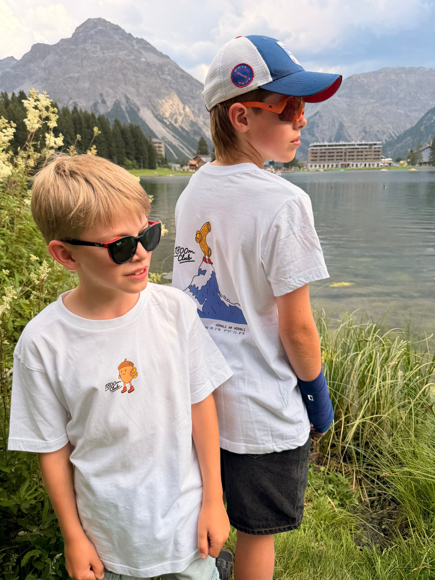 Two boys wearing white graphic t-shirts stand outdoors by the Arosa Obersee lake with mountains in the background. The boy on the right wears a blue and white cap and sunglasses, while the boy on the left wears black sunglasses.