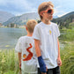 Two boys outdoors near the Arosa Obersee Lake and mountains, wearing white graphic T-shirts. One shirt has a small yellow pasta graphic, and the other has a large red squirrel path graphic.
