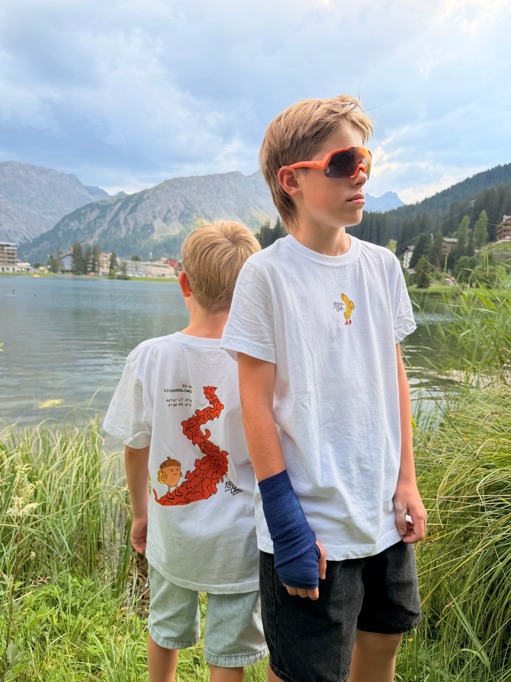 Two boys outdoors near the Arosa Obersee Lake and mountains, wearing white graphic T-shirts. One shirt has a small yellow pasta graphic, and the other has a large red squirrel path graphic.