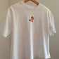 A white short-sleeve crewneck t-shirt hanging on a wooden hanger against a beige wall. The shirt features a small graphic print in the middle of a nut from the Eichhönrliweg Trail Series. 