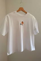 A white short-sleeve crewneck t-shirt hanging on a wooden hanger against a beige wall. The shirt features a small graphic print in the middle of a nut from the Eichhönrliweg Trail Series. 