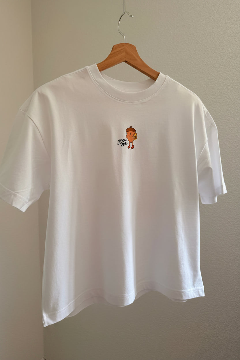 A white short-sleeve crewneck t-shirt hanging on a wooden hanger against a beige wall. The shirt features a small graphic print in the middle of a nut from the Eichhönrliweg Trail Series. 