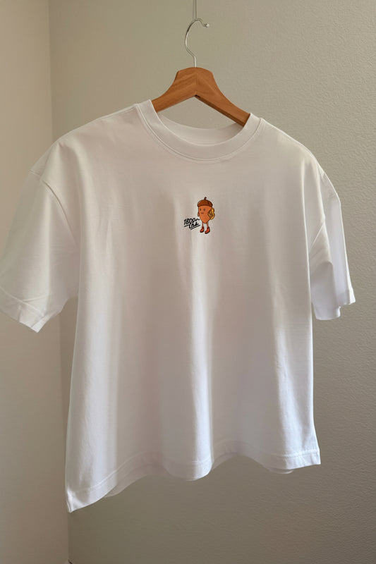 A white short-sleeve crewneck t-shirt hanging on a wooden hanger against a beige wall. The shirt features a small graphic print in the middle of a nut from the Eichhönrliweg Trail Series. 