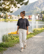 A woman walking along the Arosa Obersee wearing the 1800m Club Hörnli embroidered Boxy T-shirt designed in Arosa, Switzerland.