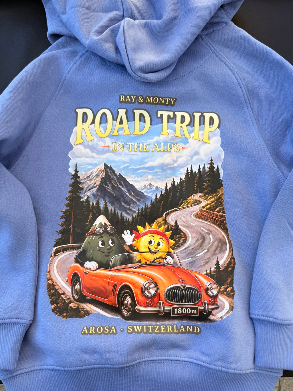 Close-up of the Road Trip in the Alps back print, with Ray and Monty in a red convertible on a winding mountain road.