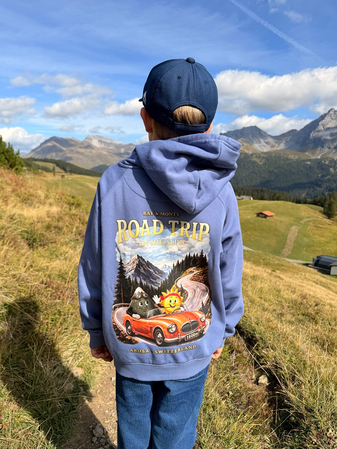 Back of the blue Ray & Monty Road Trip kids' hoodie, showing the large Alpine road-trip illustration on a mountain path.