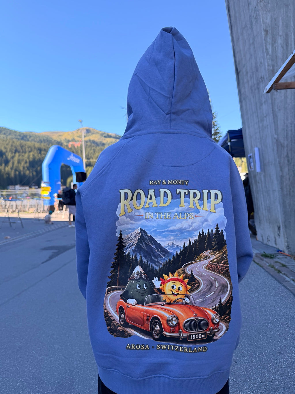 Ray & Monty Road Trip kids' hoodie worn outdoors with the hood up, showing the red convertible and Alpine road back print.